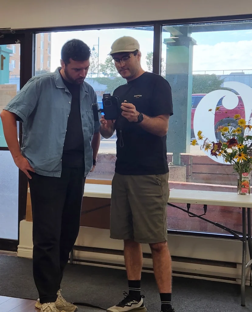 Deux hommes examinent l'écran d'une caméra de tournage devant la vitrine de l'Outillerie Rimouski.